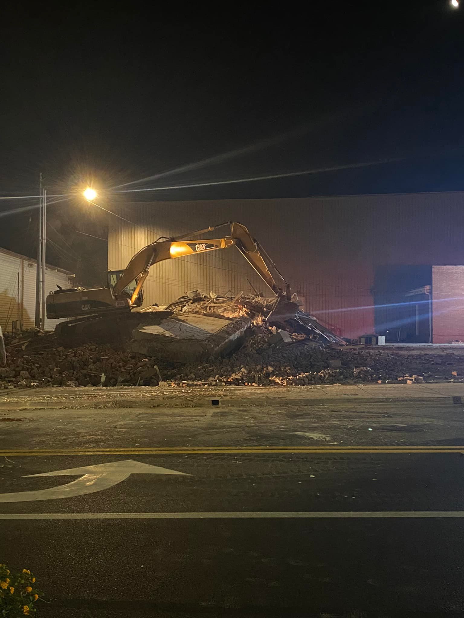 A yellow excavator works on a pile of rubble and debris in front of a building at night.
