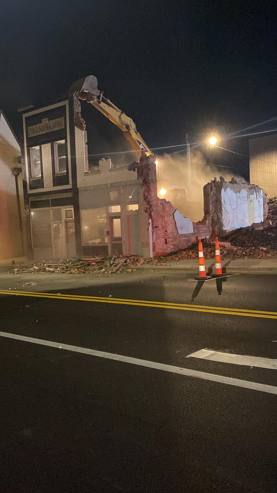 An excavator demolishes a building at night, creating dust near a road with orange traffic cones.