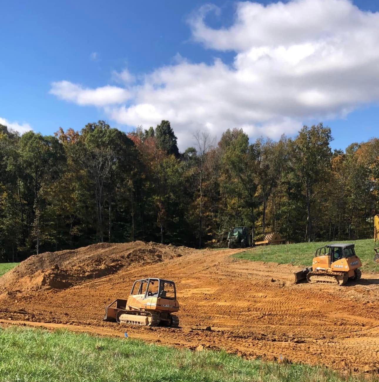 Two yellow bulldozers clear dirt on a large, rural construction site near a dense treeline under a bright blue sky.