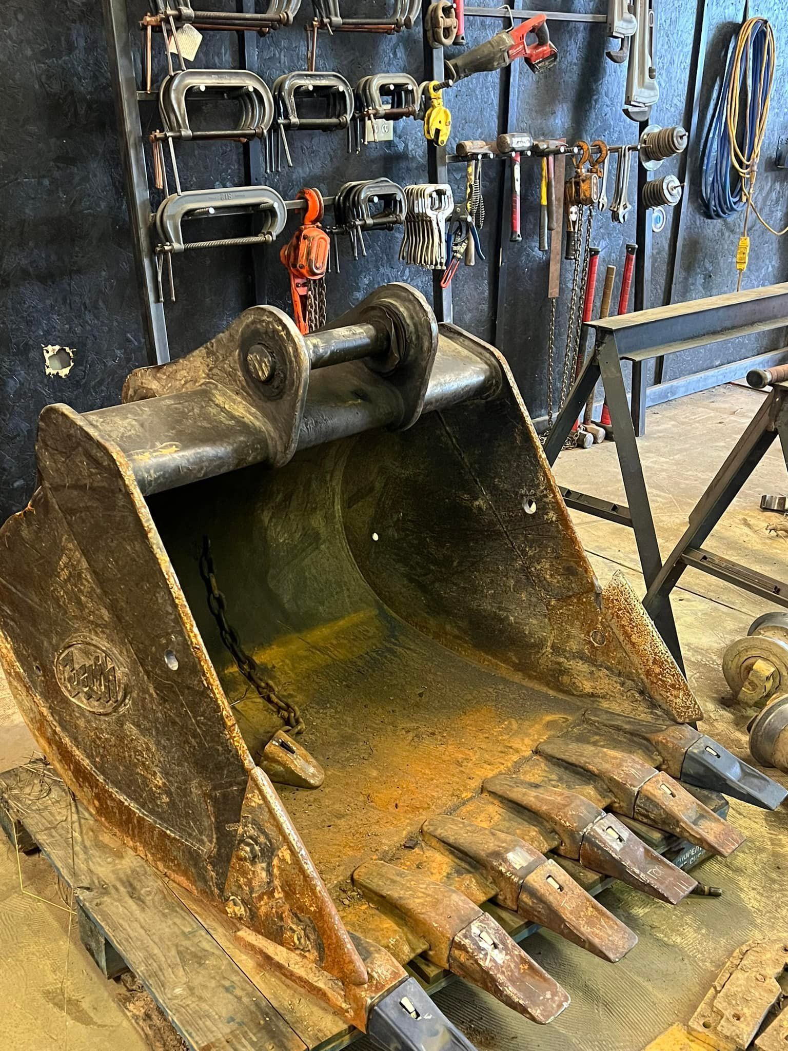 A metal excavator bucket sits on a wooden pallet inside a workshop with various tools hanging on the wall behind it.