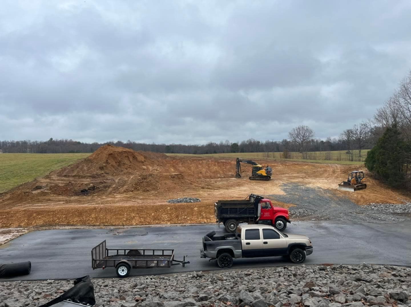 A construction site featuring an excavator, a red dump truck, and a pickup truck towing a trailer on a paved lot.