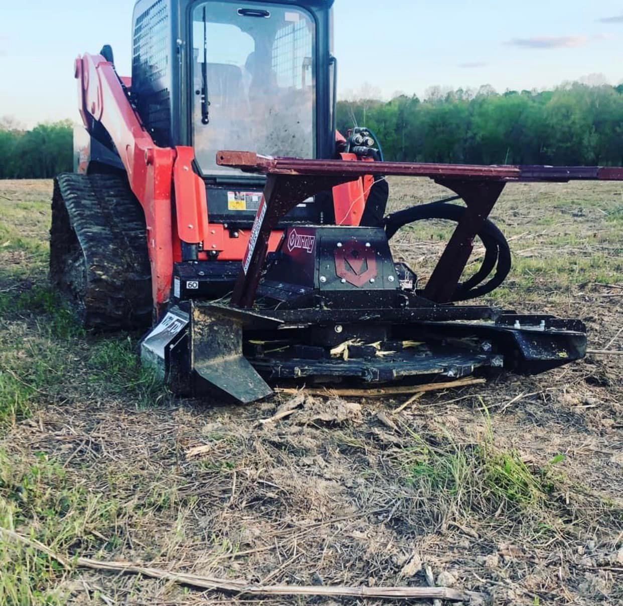 An orange skid steer loader with a heavy-duty brush cutter attachment sits on a dirt field with trees in the background.
