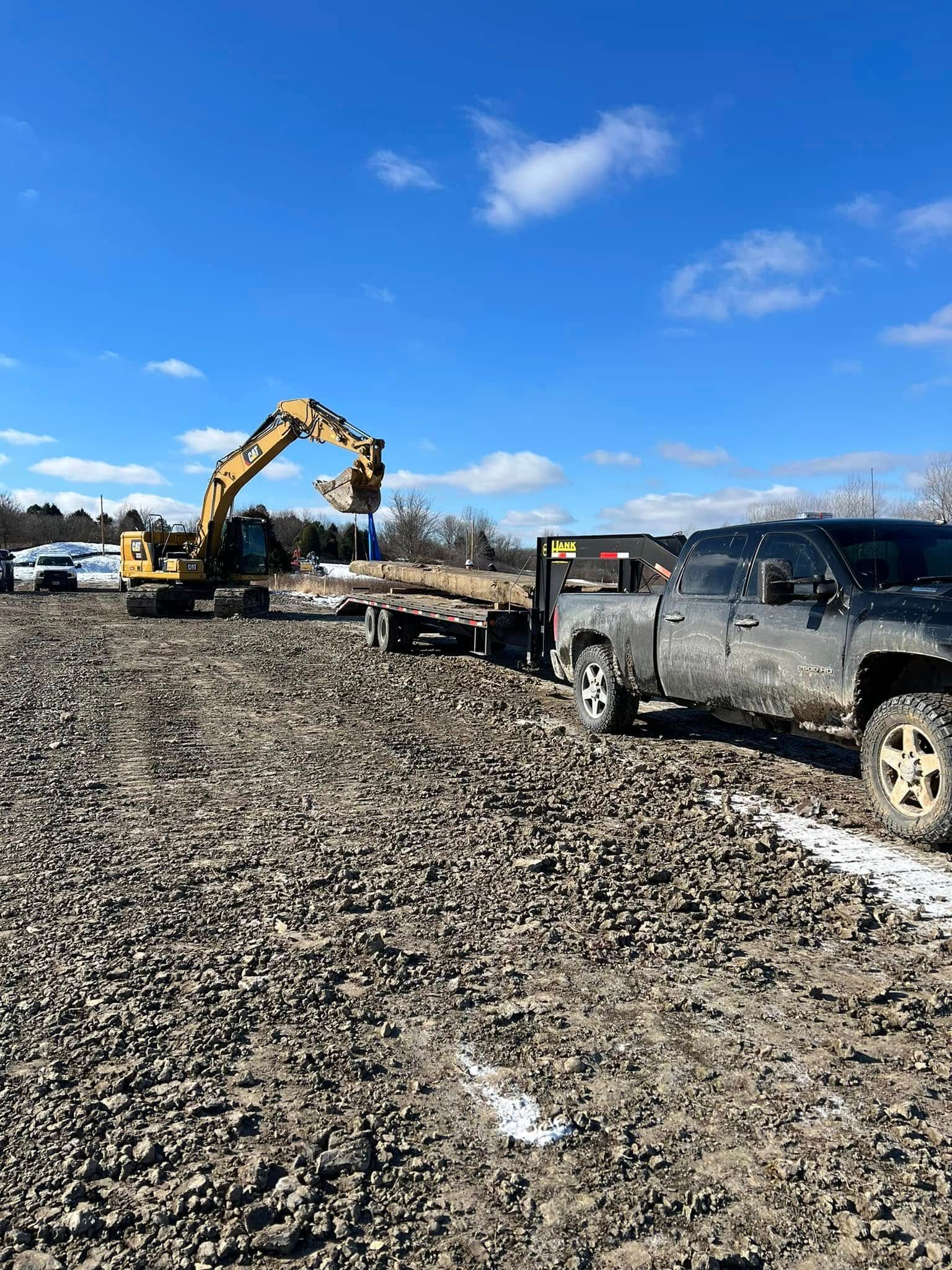 An excavator with a hydraulic breaker works on a gravel lot next to a gray pickup truck under a clear blue sky.