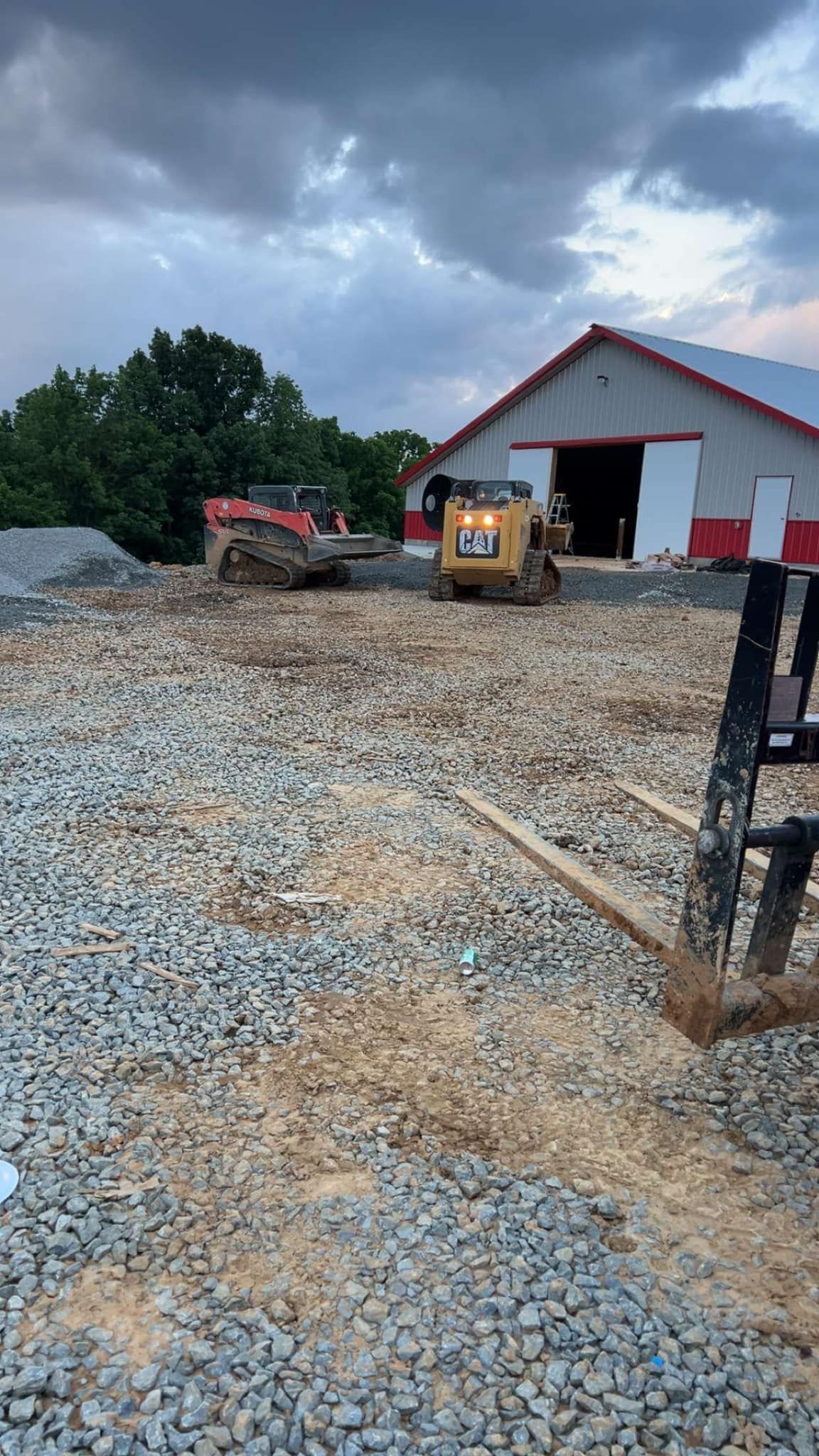 Two skid-steer loaders sit on a gravel and wood chip construction site in front of a white barn under a stormy sky.