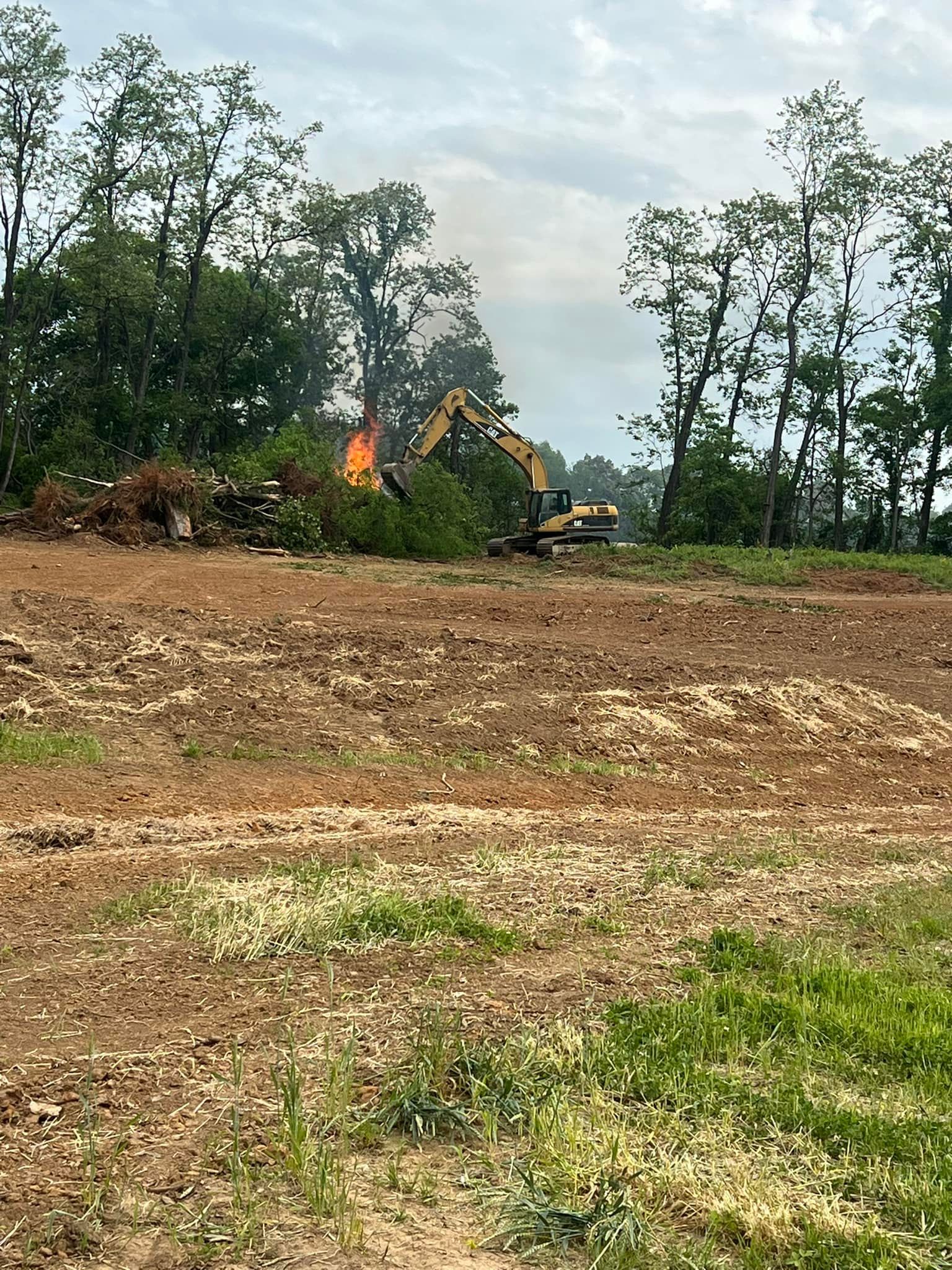A yellow excavator works near a pile of brush that is actively burning in a cleared field bordered by trees.