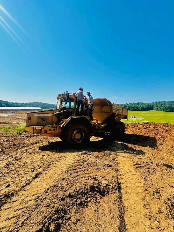 Two people stand atop a yellow construction dump truck on a dirt site under a clear blue sky.