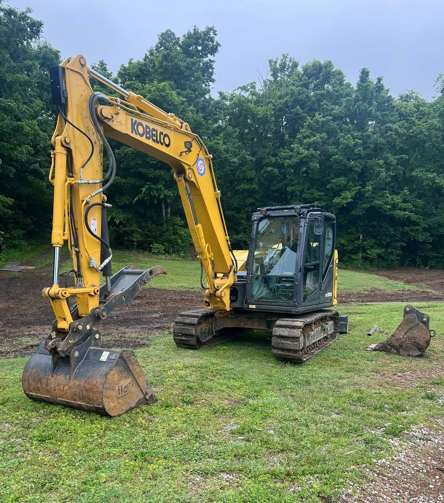 A white and red excavator digging a trench in a wooded area, with a long, black corrugated drainage pipe being installed.
