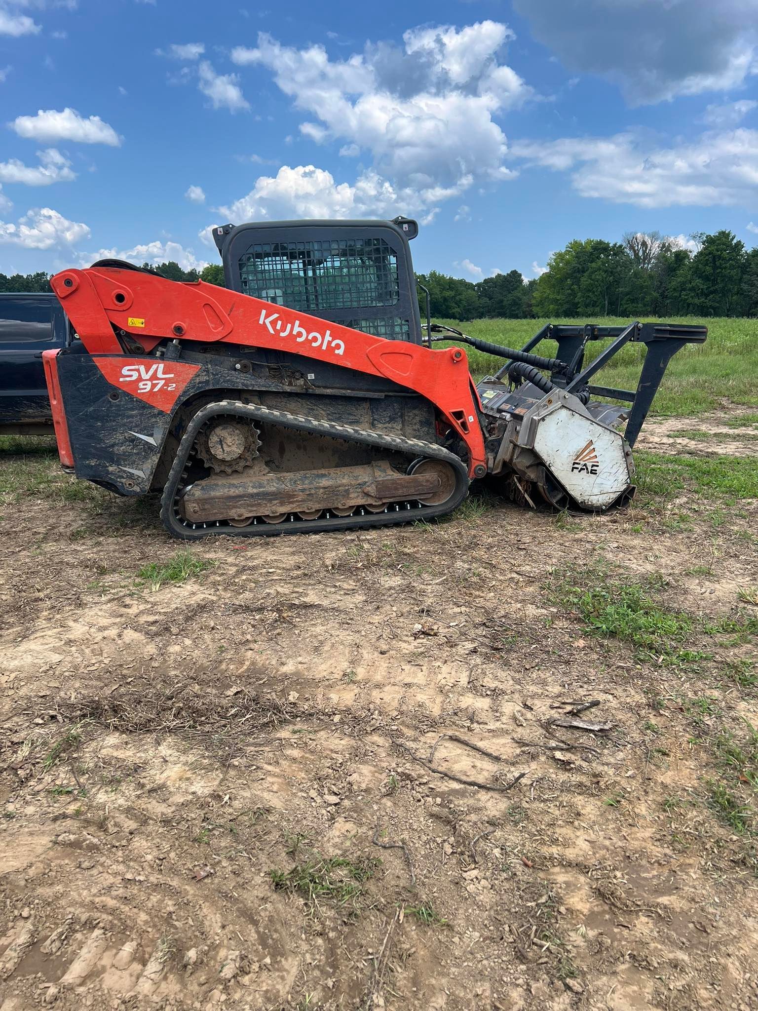 A bright orange Kubota compact track loader with a forestry mulcher attachment sits in a dirt field under a cloudy sky.