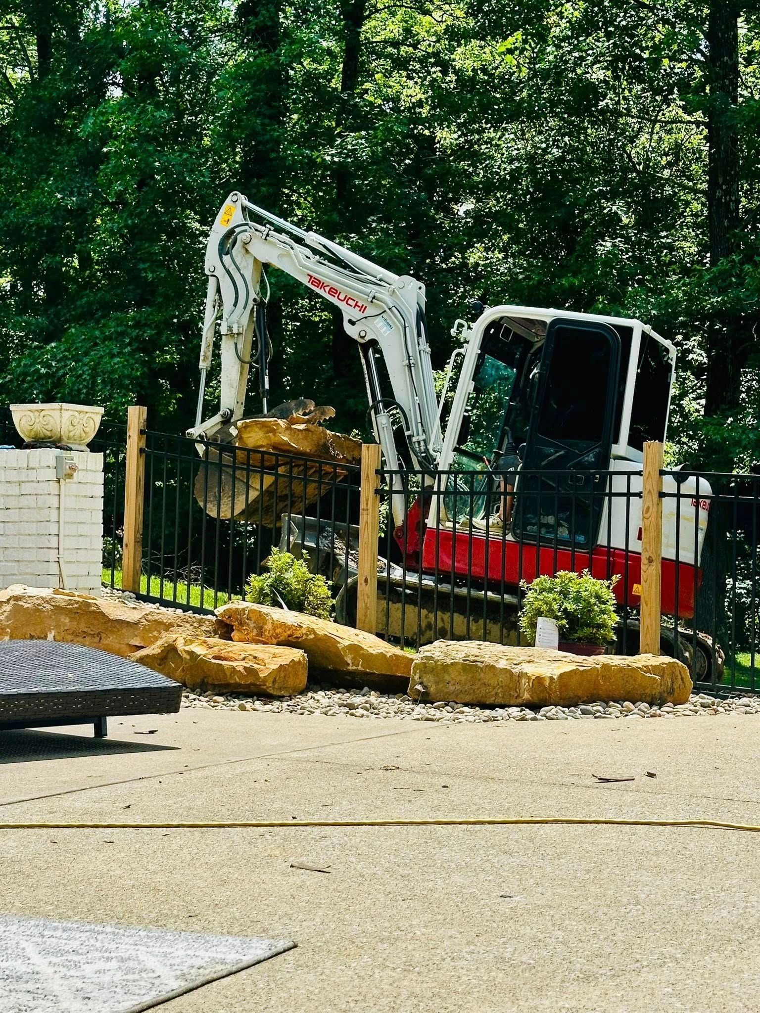 A white construction excavator sits behind a black fence, lifting large yellow rocks in a sunny, tree-lined yard.