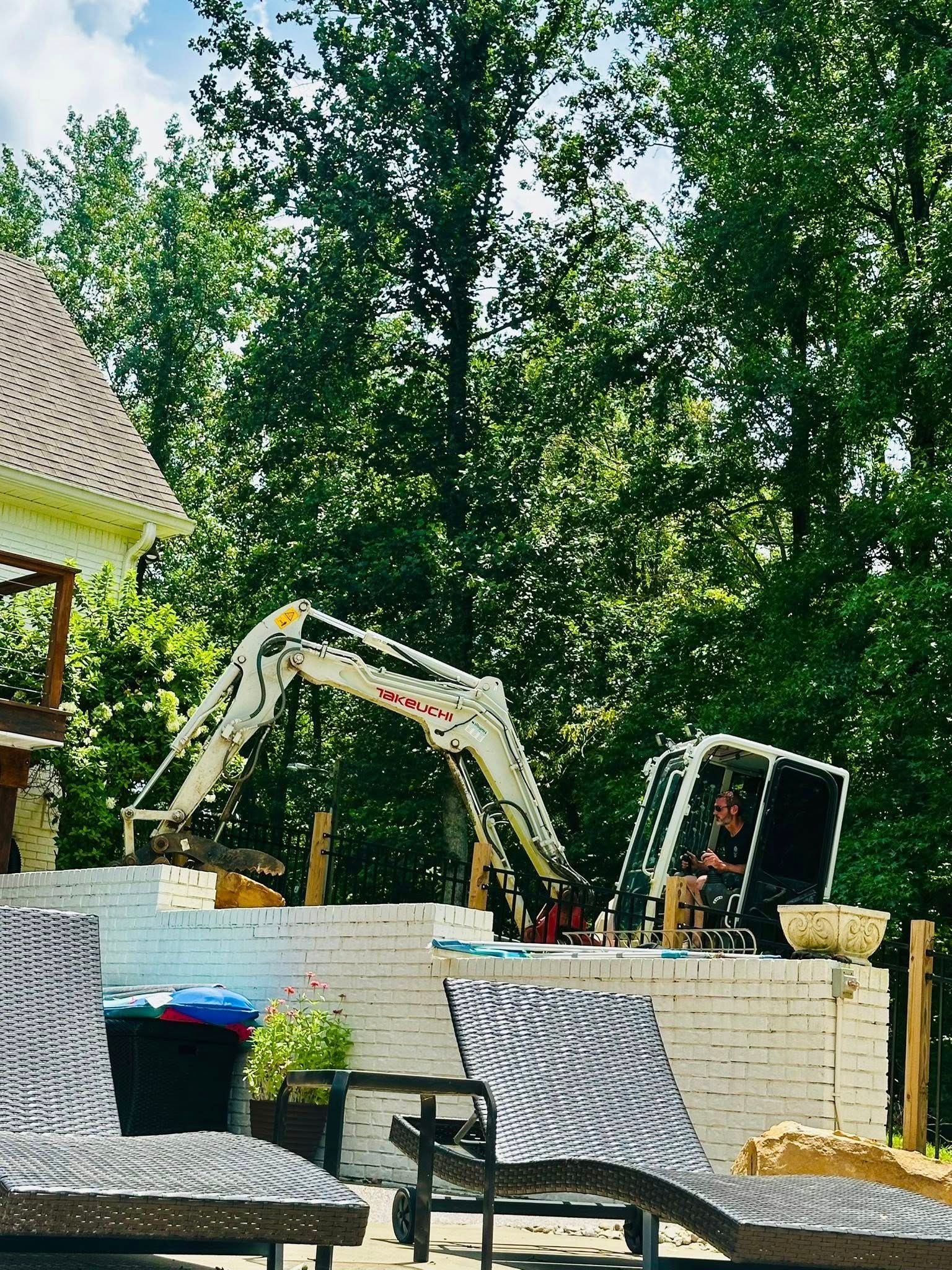A small white excavator operates behind a stone wall near a patio with lounge chairs and green trees in the background.