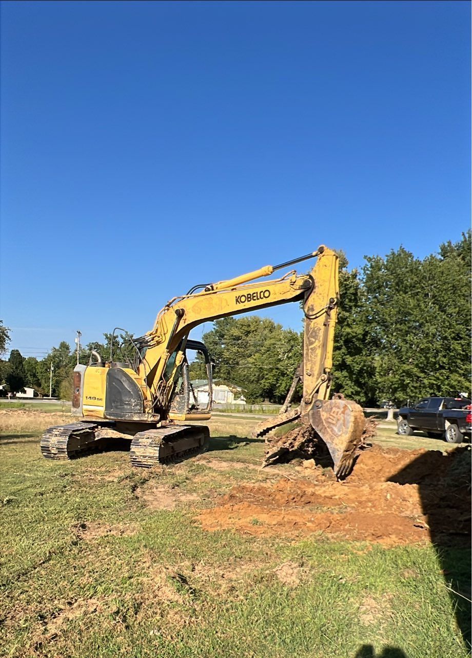 A yellow Case excavator sits on a grassy lot, its digging bucket lowered toward a patch of exposed dirt under a blue sky.