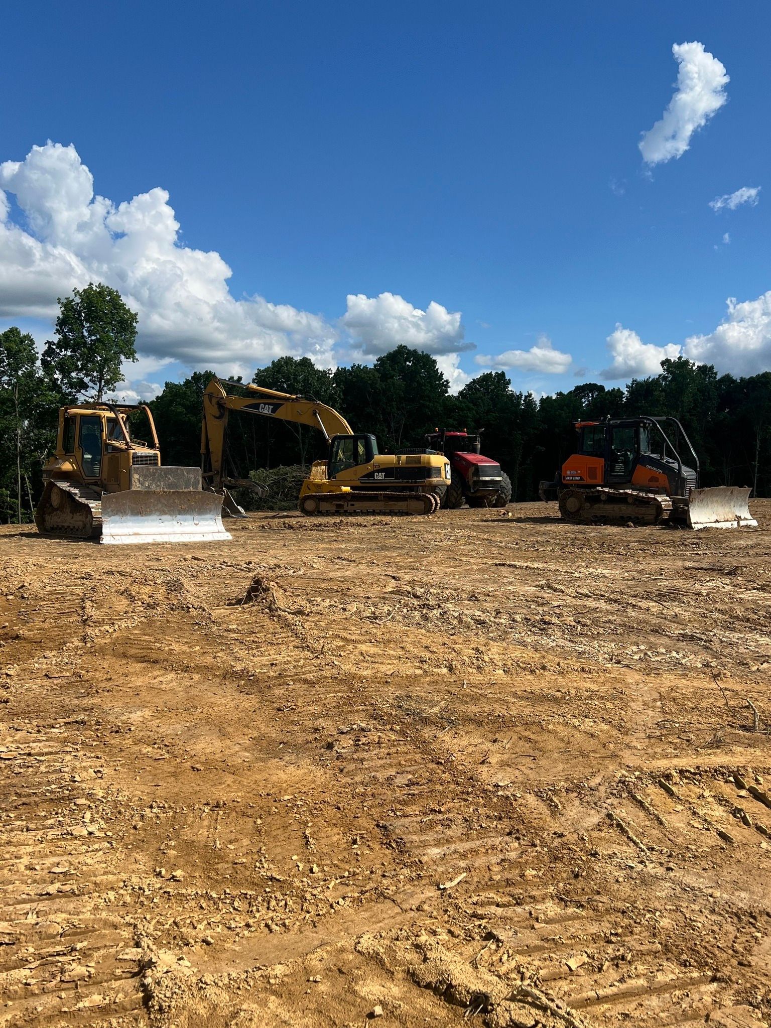Several pieces of heavy construction machinery, including excavators and bulldozers, sit on a dirt site under a blue sky.