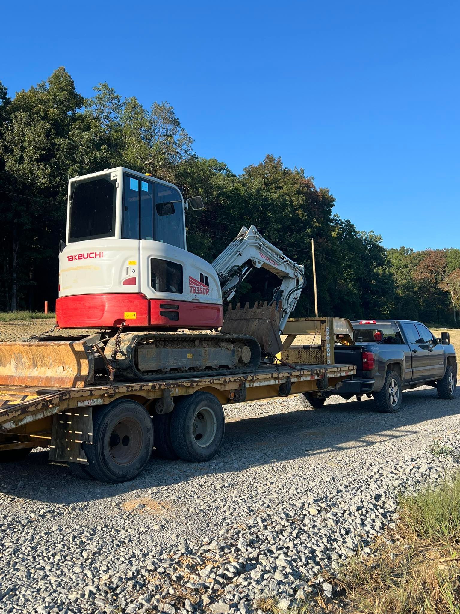 A white and red excavator loaded on a flatbed trailer being towed by a pickup truck on a gravel road by a tree line.
