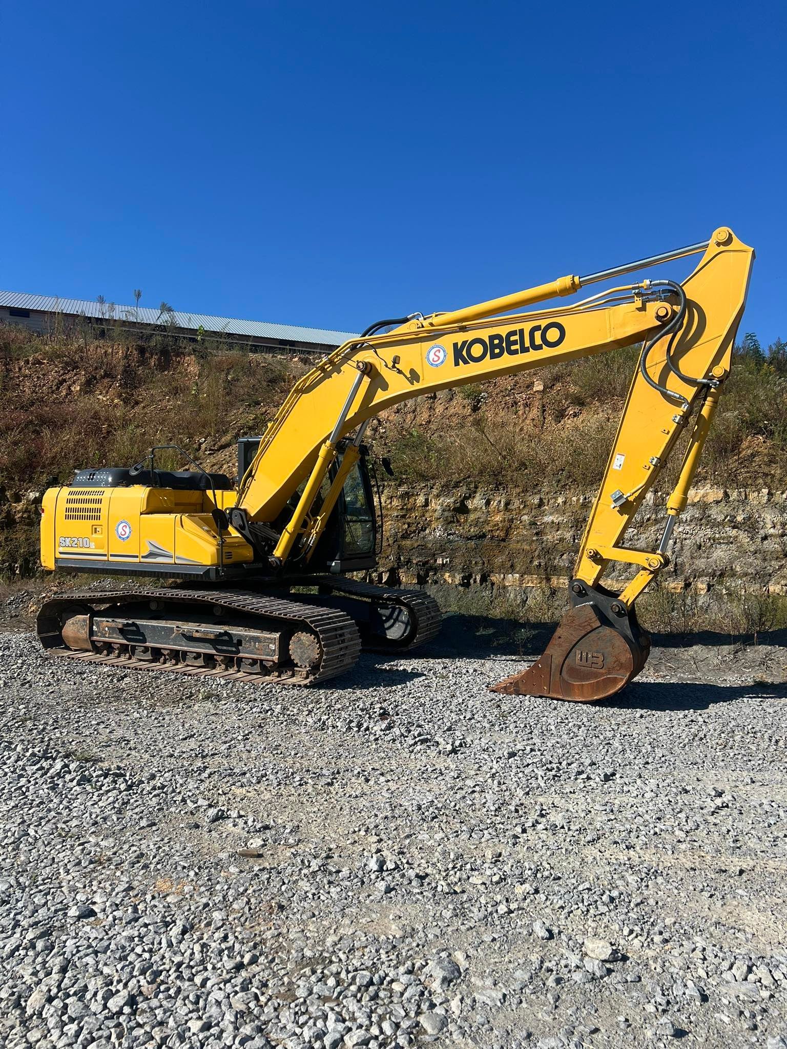 A yellow Kobelco excavator parked on a gravel surface under a clear blue sky.
