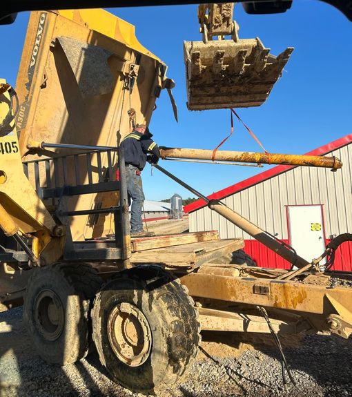 A worker in a high-visibility jacket stands on a yellow construction vehicle while holding a long, suspended pipe.