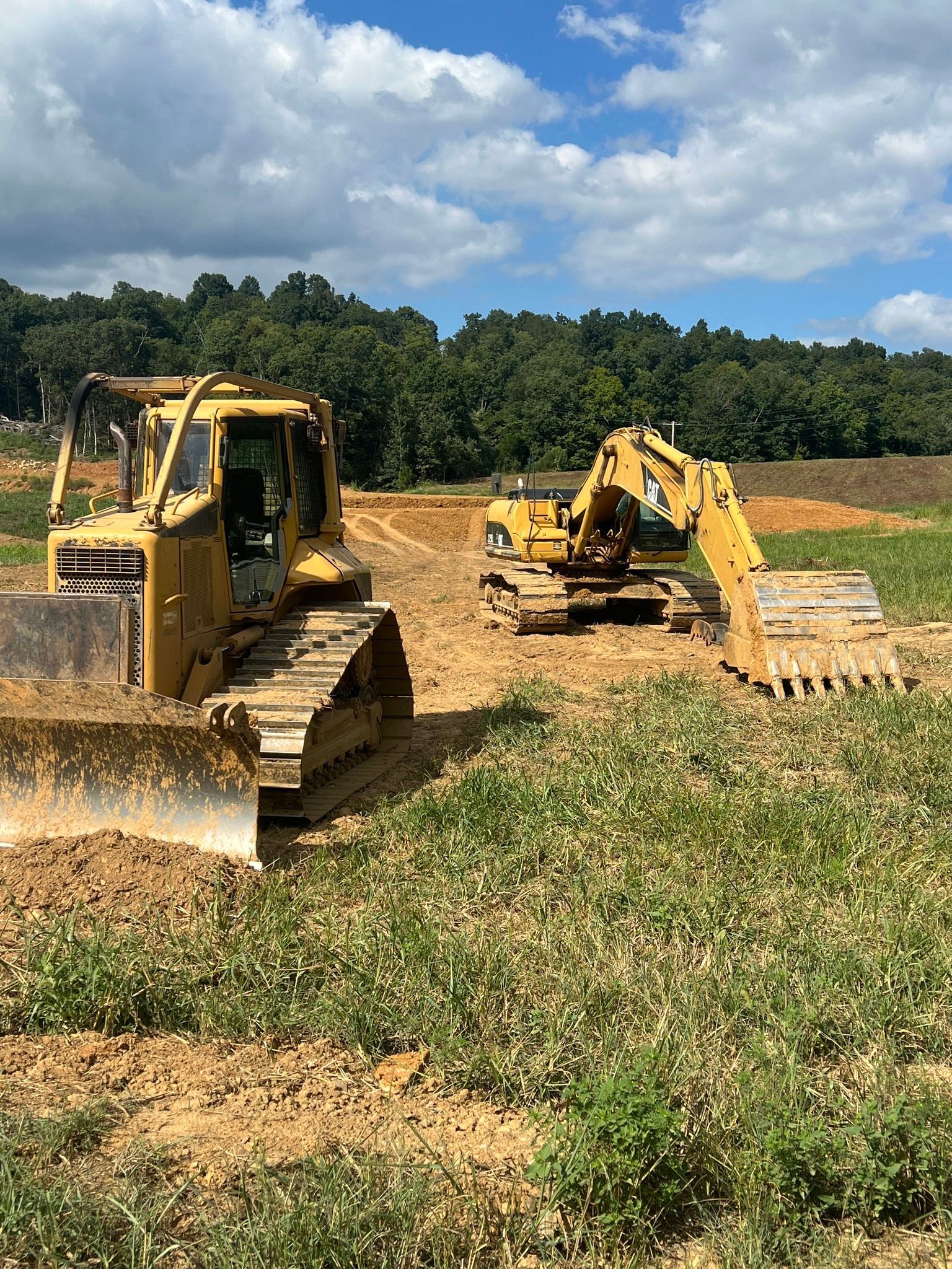 Two yellow construction machines sit on a dirt lot in front of a treeline under a blue, cloudy sky.
