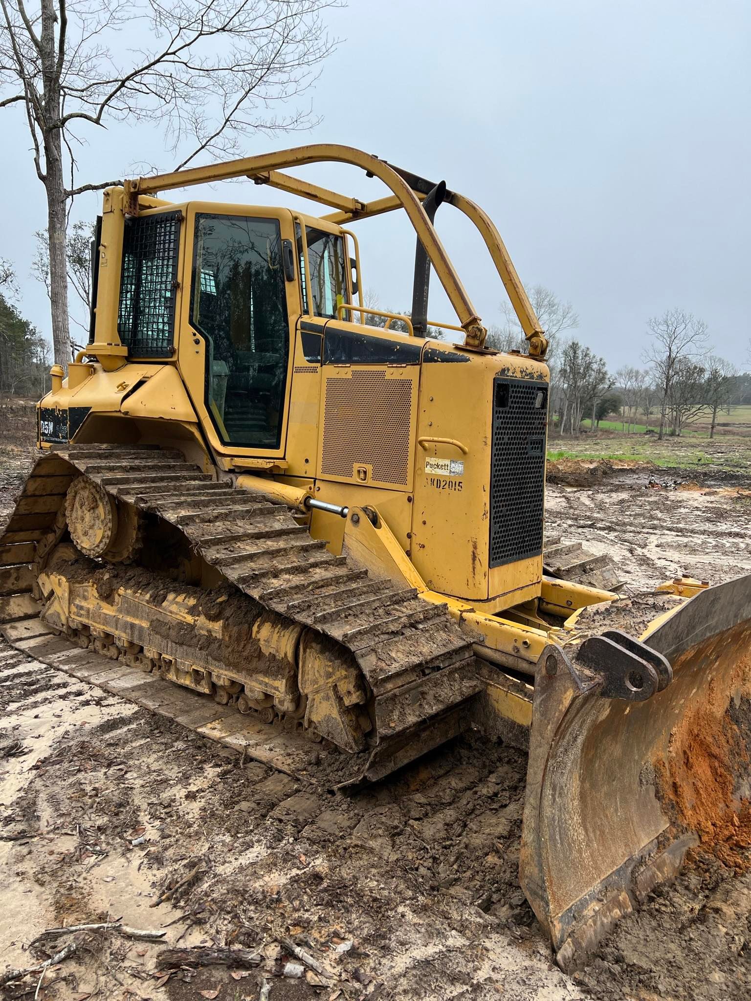 A yellow bulldozer with tracks sits on muddy ground in a clearing surrounded by trees.