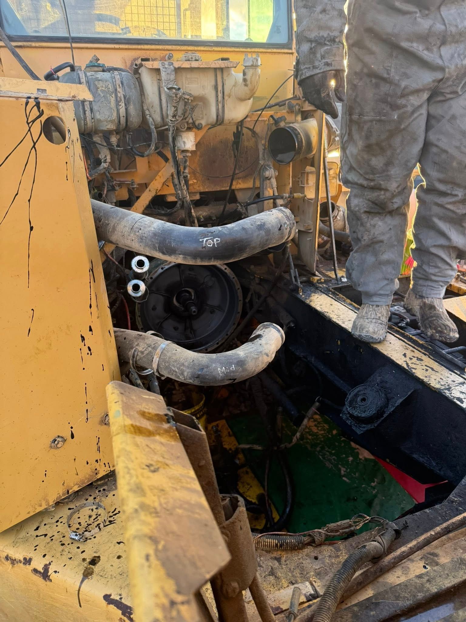 A mechanic in dirty coveralls stands on a yellow industrial machine, repairing a large, exposed engine assembly.