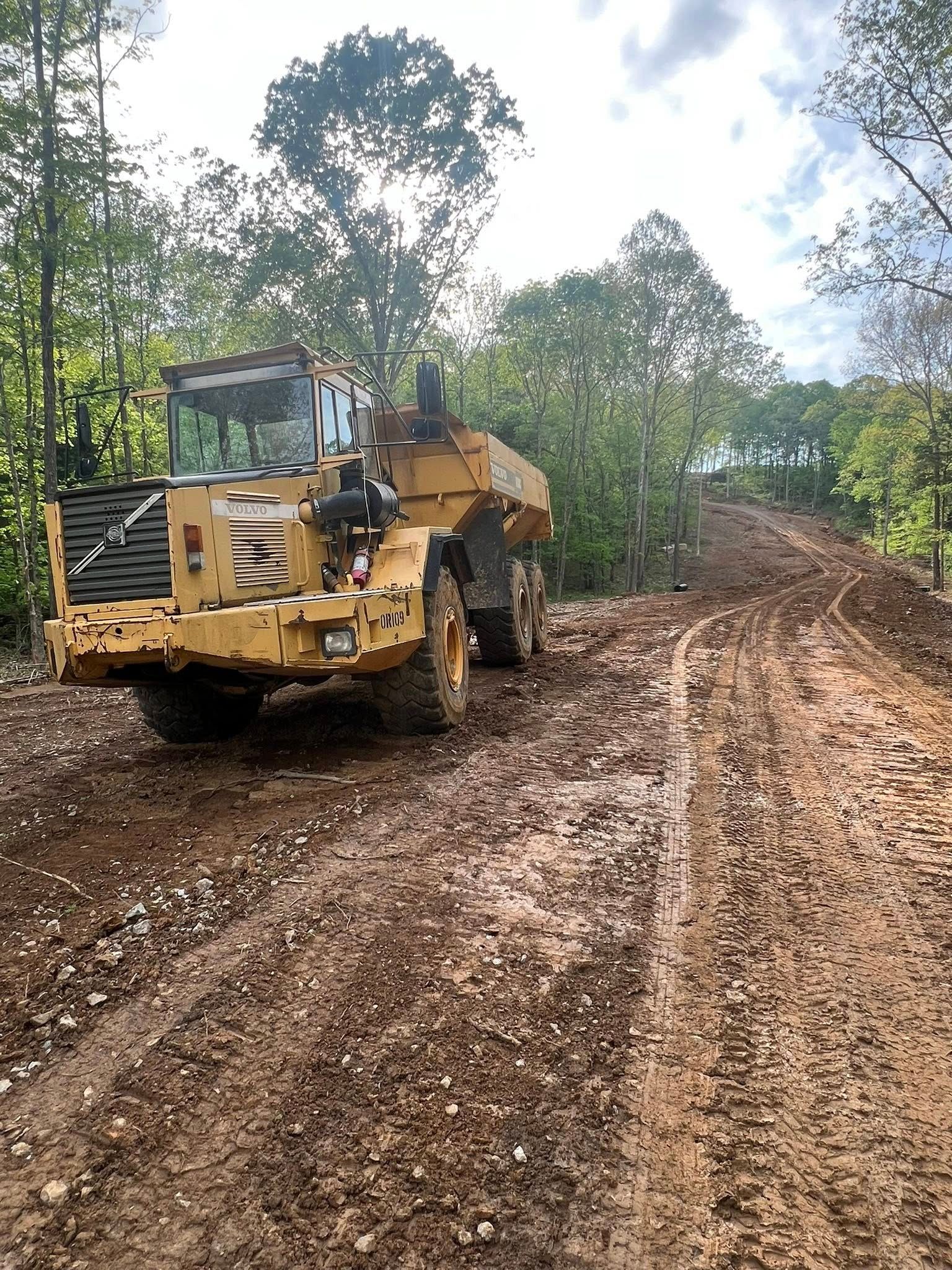 A yellow Volvo articulated hauler parked on a muddy, dirt construction path through a forest.