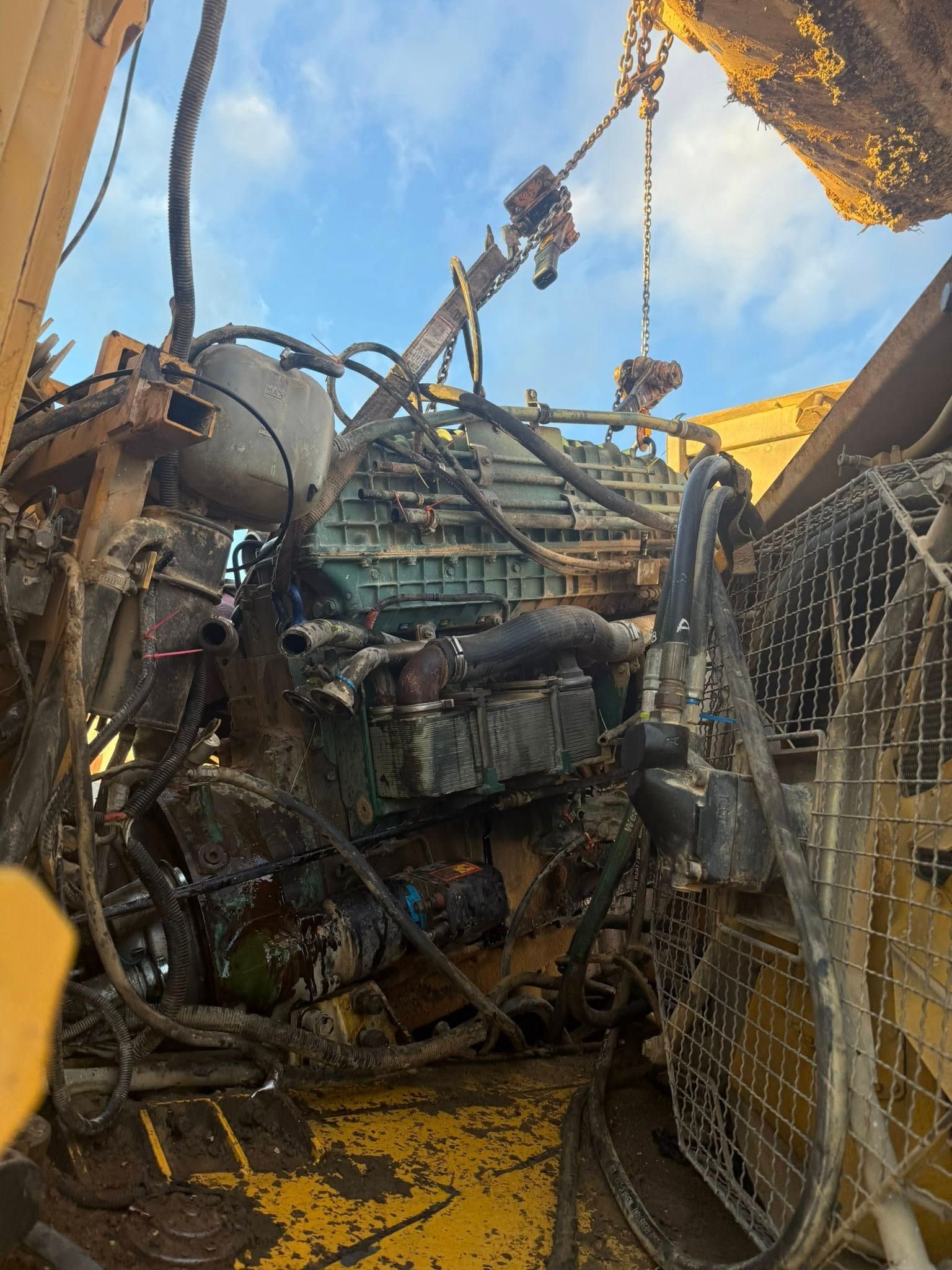 A close-up view of a large, weathered industrial engine with wires and hoses, being lifted outdoors by a crane.