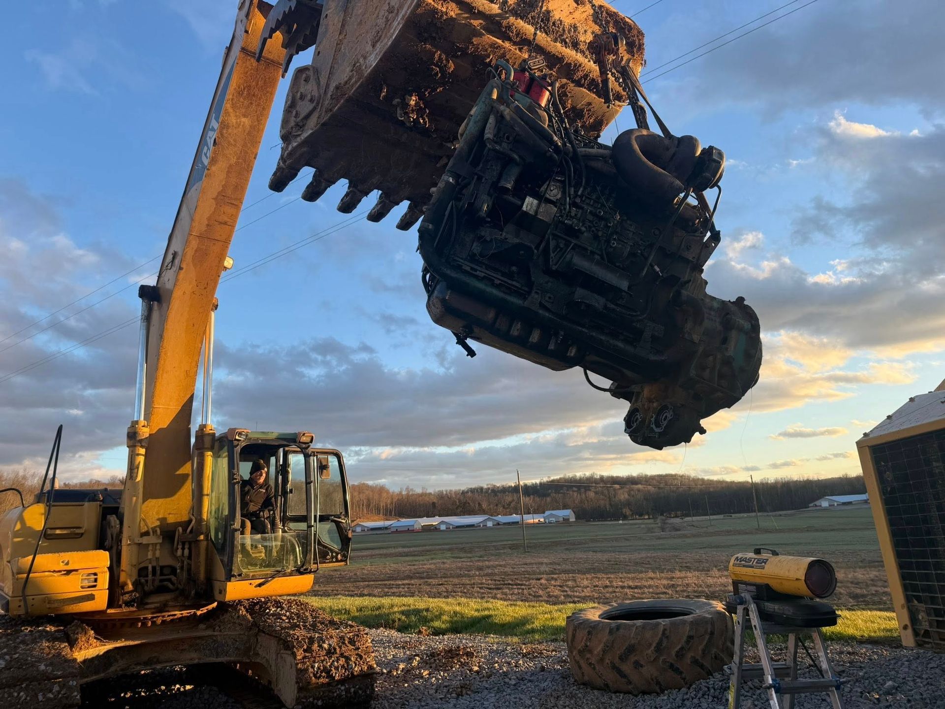 A yellow excavator lifting a large, dark engine with its bucket in an open field at sunset.