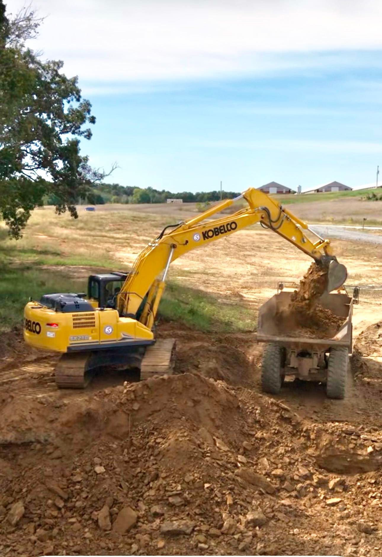 A yellow Kobelco excavator filling a dump truck with soil at a construction site.