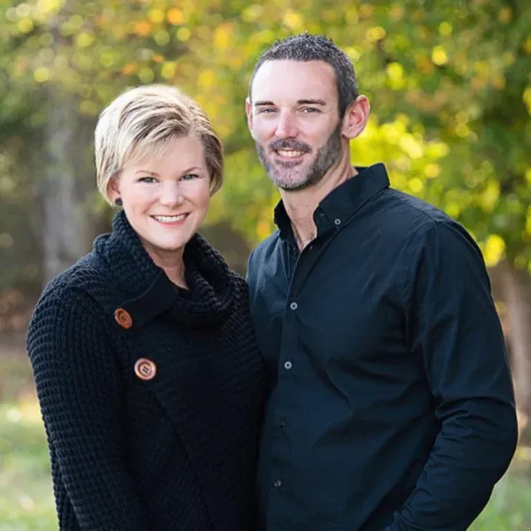 A couple posing for a portrait in a park, both wearing black clothing, with a blurred green background.