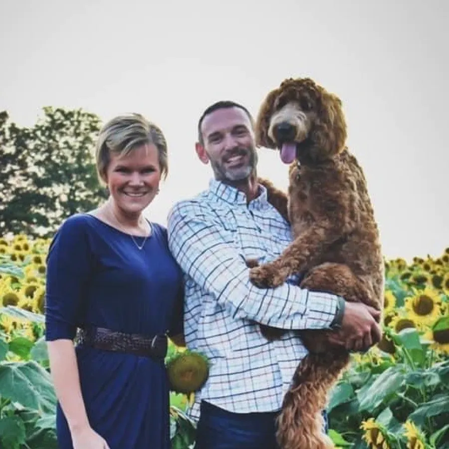 A person in a blue dress and another in a plaid shirt hold a brown curly-haired dog in a field of sunflowers.