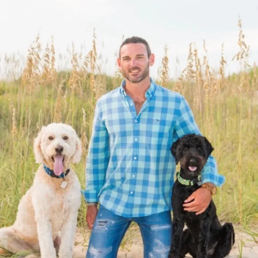 A person in a blue checkered shirt kneels in beach grass between a cream-colored dog and a black dog, smiling at the camera.
