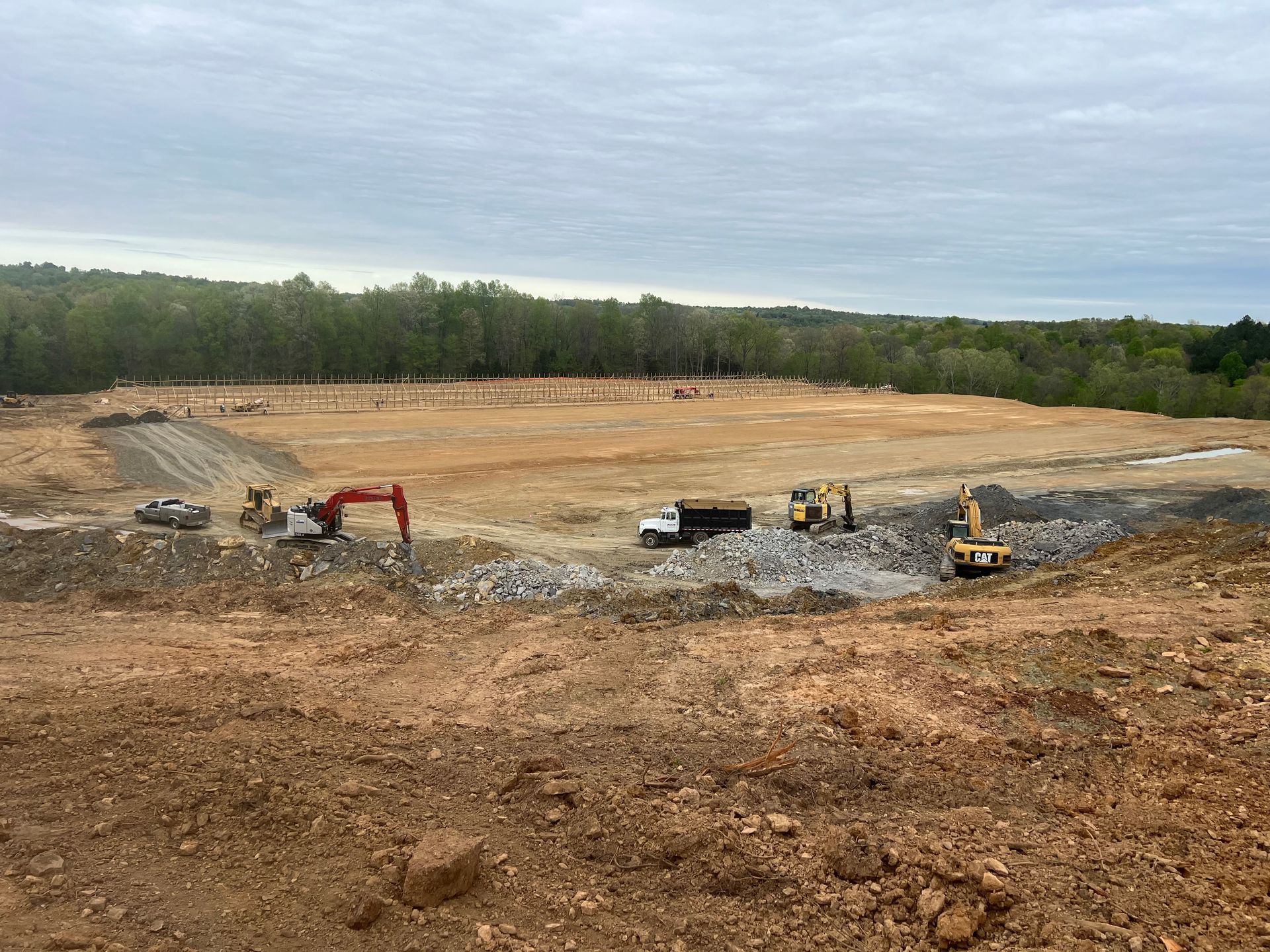 Construction site with excavators, a dump truck, and large piles of dirt and rocks under a cloudy sky.
