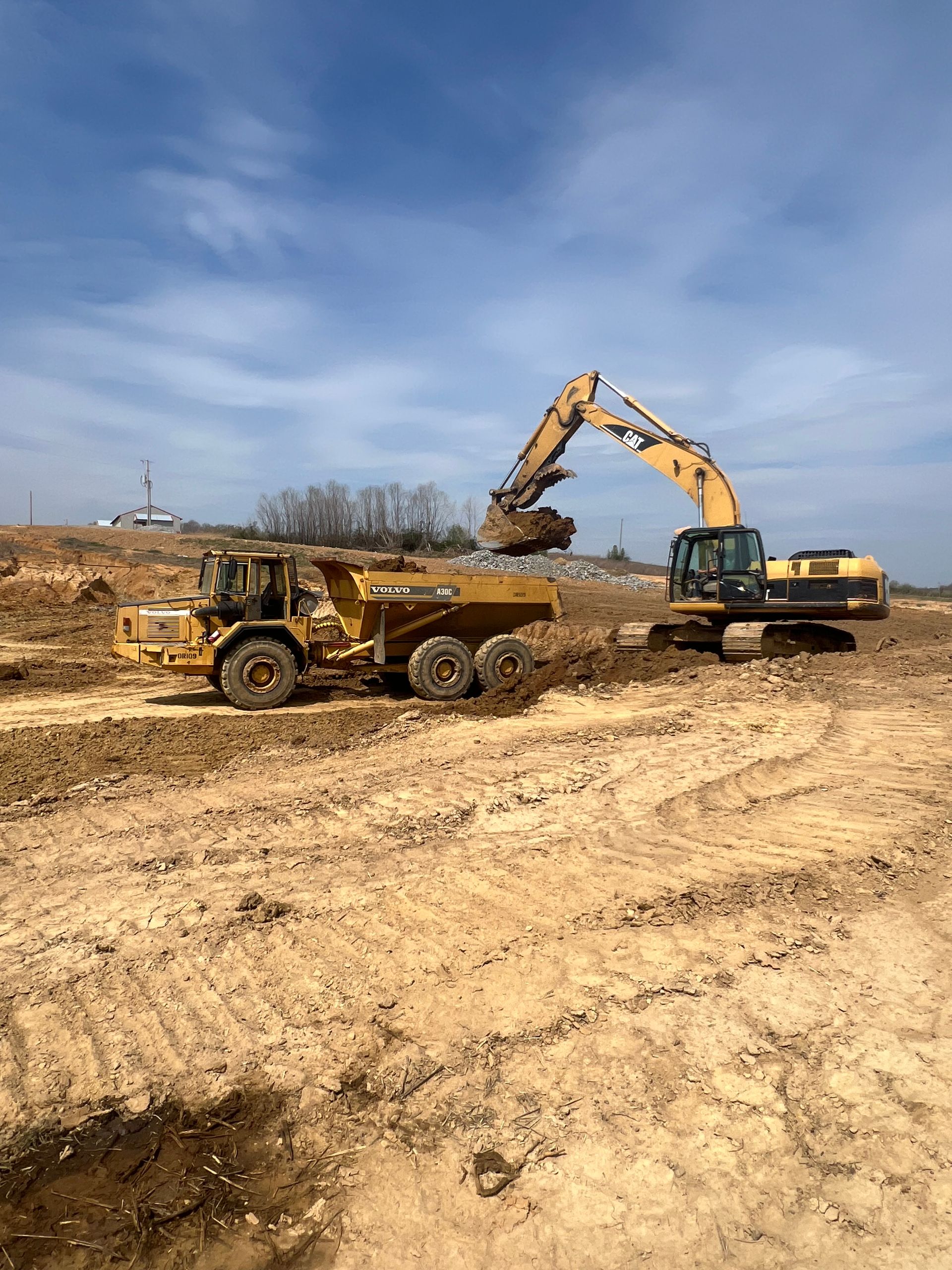 A yellow excavator fills a large, yellow articulated dump truck with dirt on a sunny construction site.