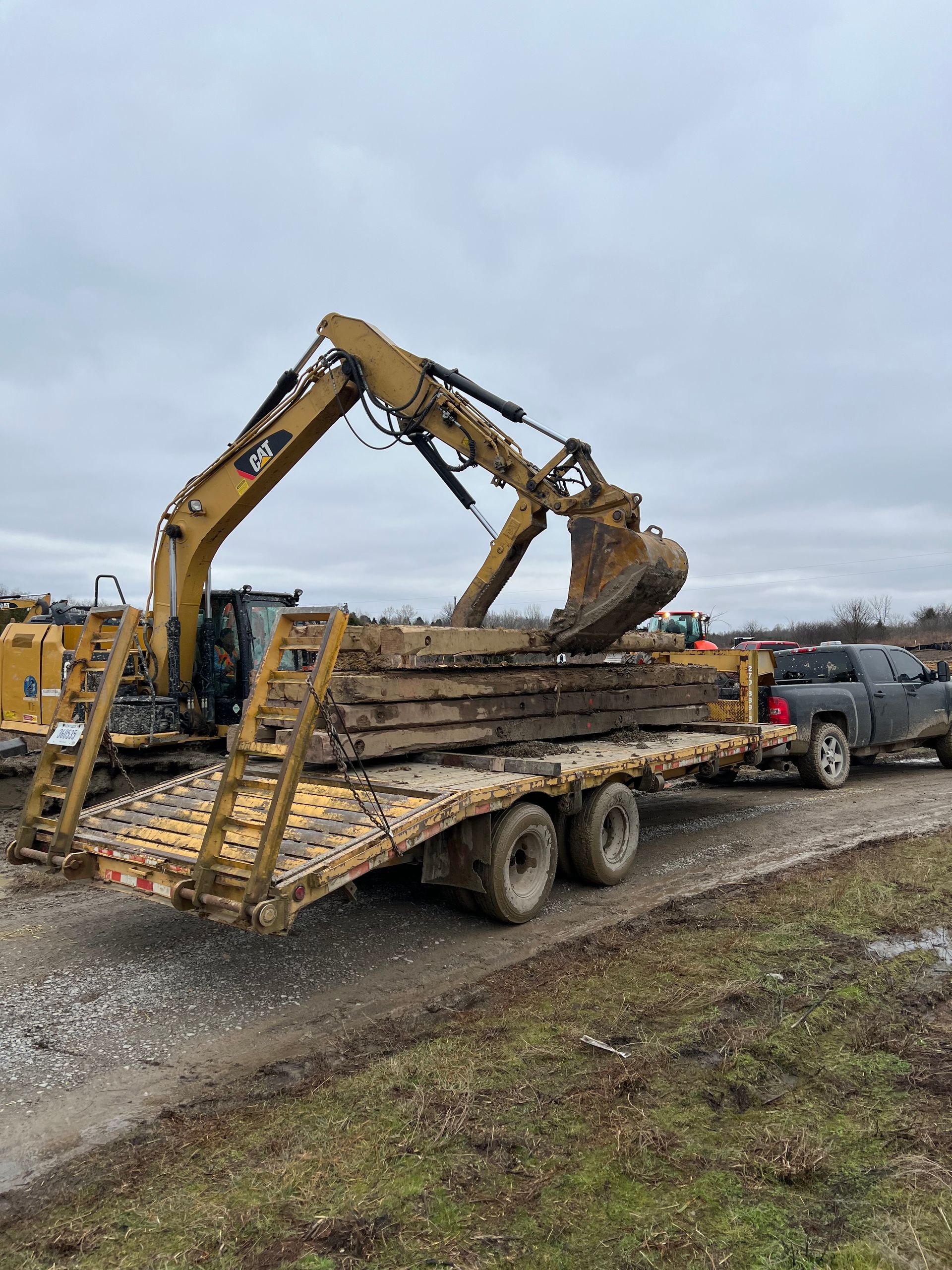 A yellow Caterpillar excavator on a flatbed trailer being towed by a gray pickup truck on a gravel road.