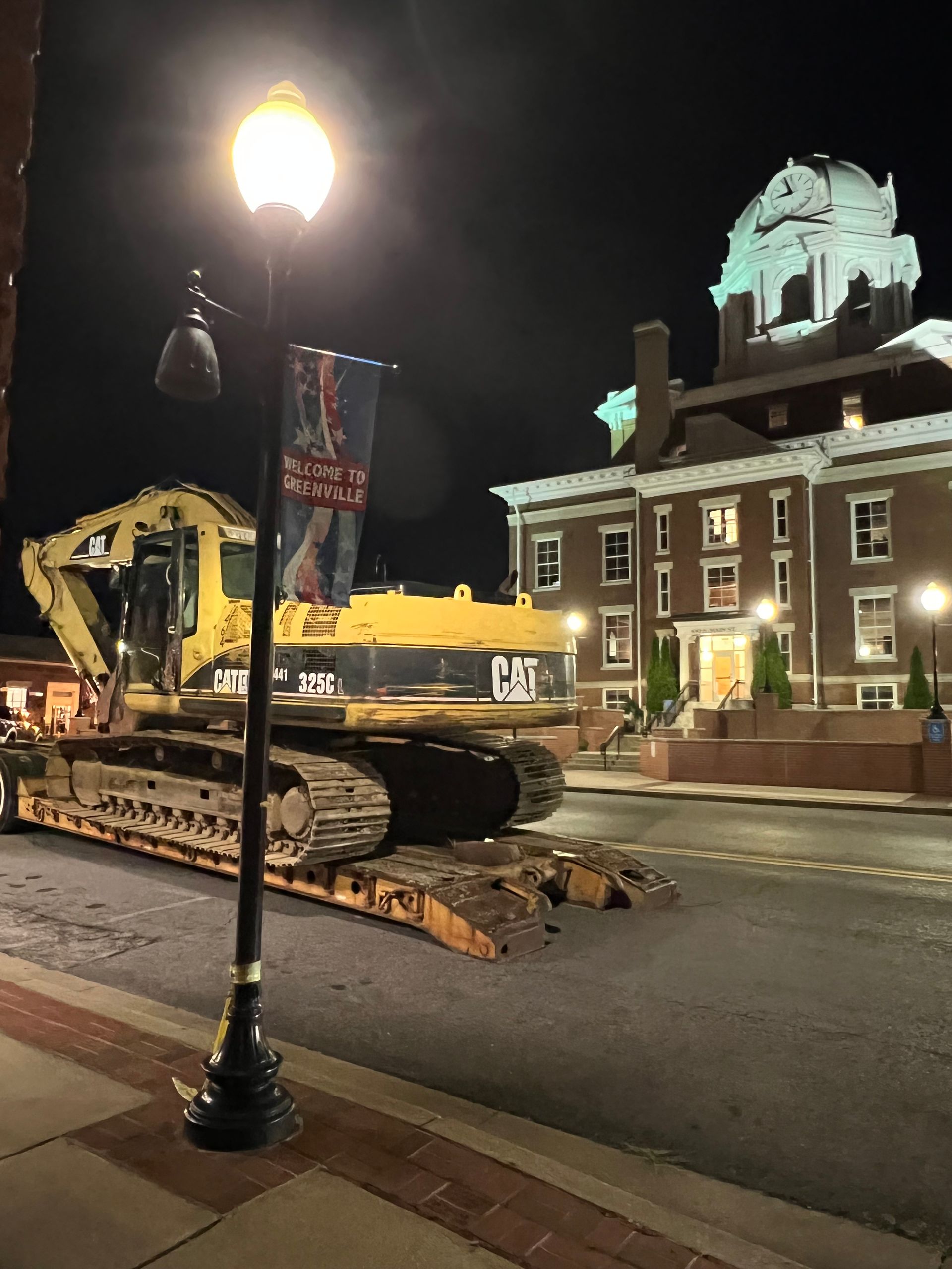 A yellow Caterpillar excavator sits on a trailer in front of a historic brick building at night under a streetlamp.