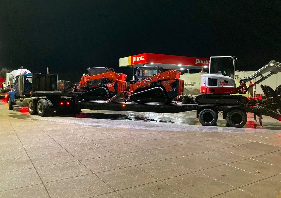 A flatbed semi-truck at a gas station at night, carrying three pieces of heavy construction equipment.