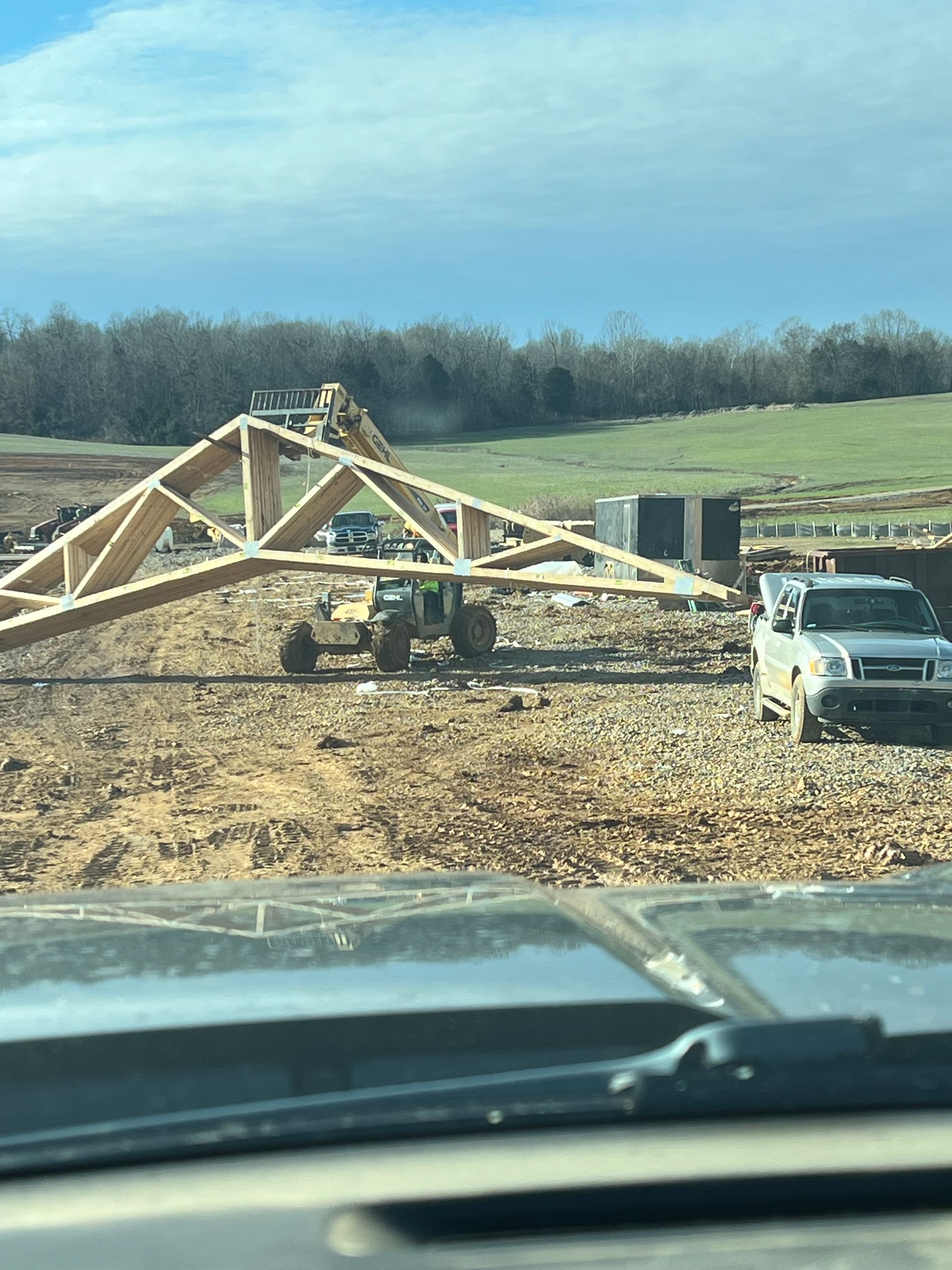 A large wooden roof truss is being lifted by a telehandler on a dirt construction site, with a pickup truck parked nearby.