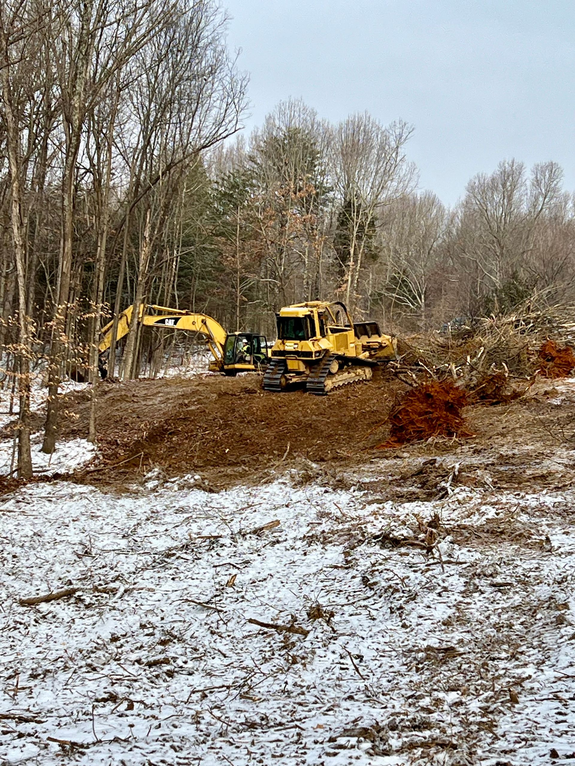 Two yellow bulldozers clear land on a partially snow-covered, wooded construction site.