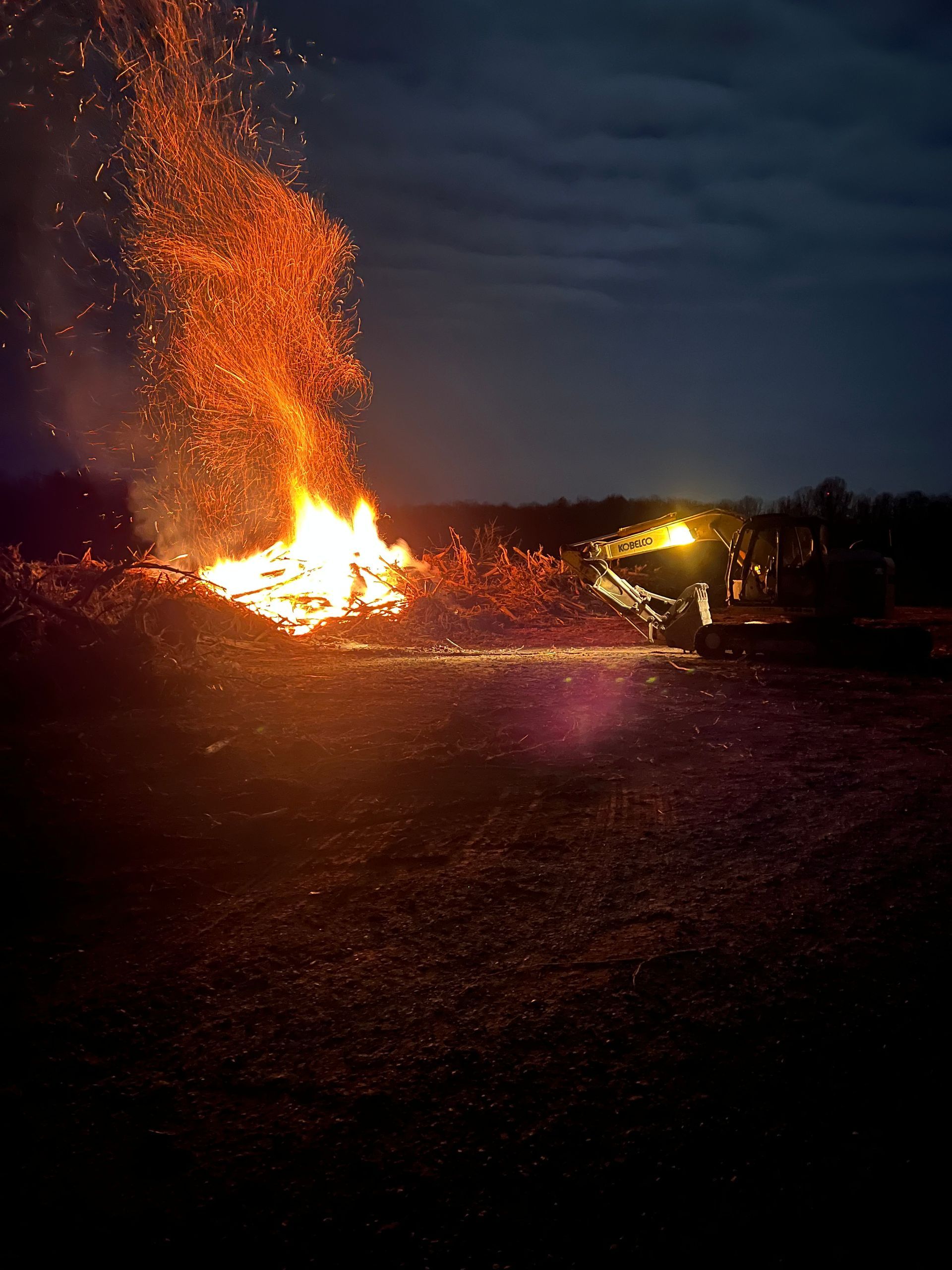 A large bonfire burns brightly in a dark field at night, with a piece of heavy machinery nearby under a cloudy sky.