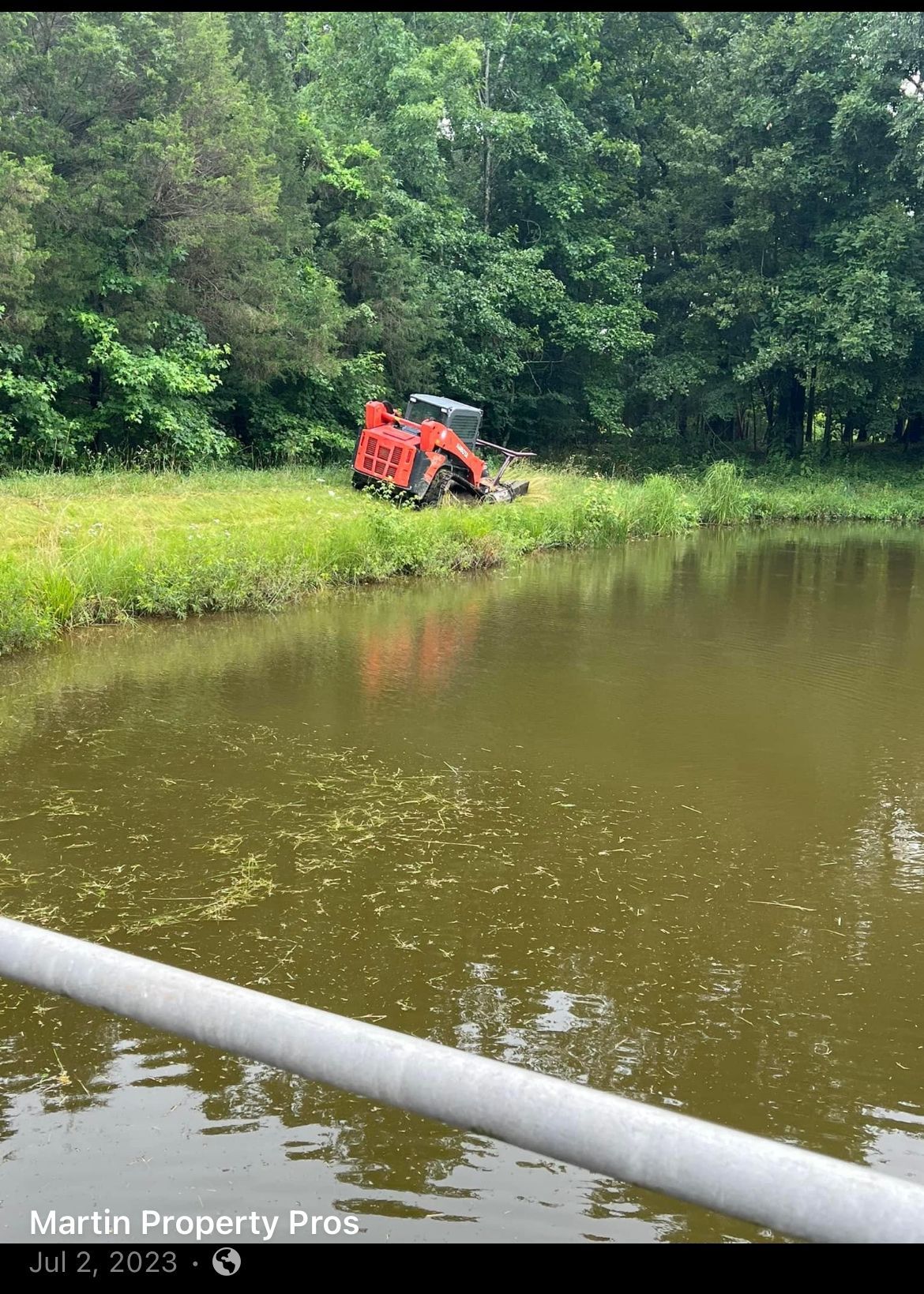 An orange skid steer mower parked on a grassy bank next to a calm pond with a white pipe in the foreground.