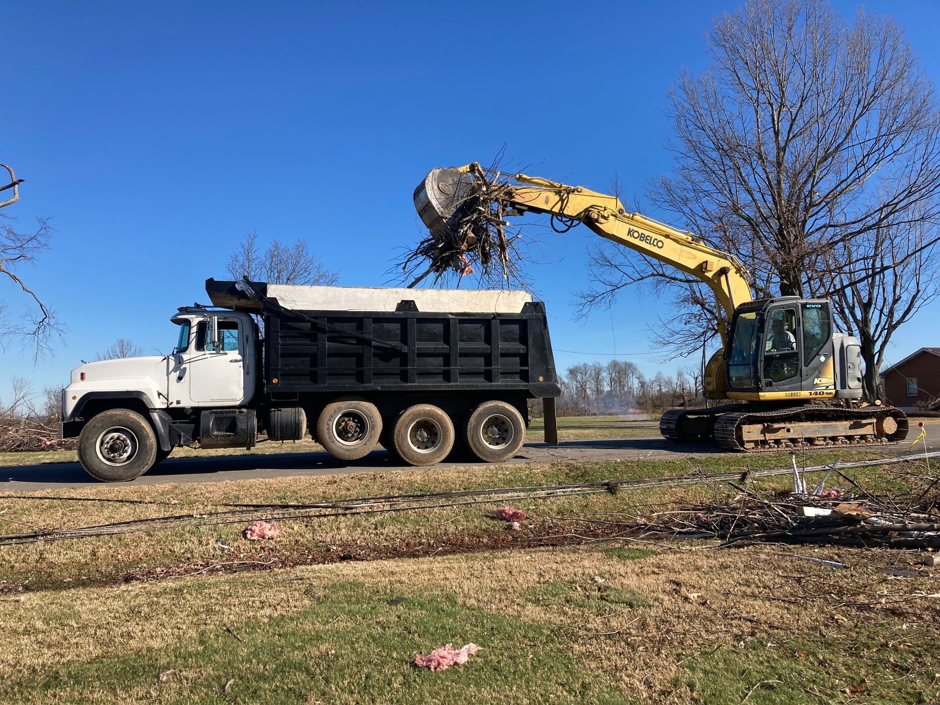 A yellow excavator loading debris into the bed of a white and black dump truck against a clear blue sky.