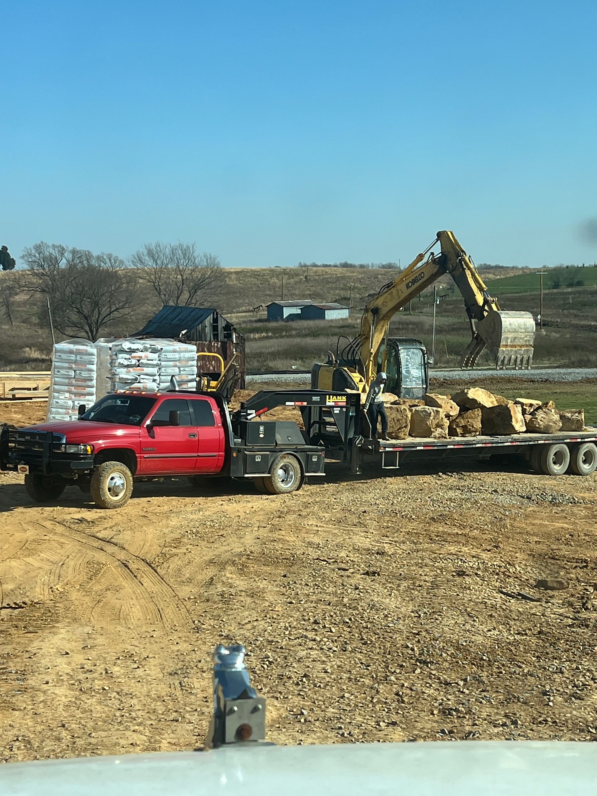 A red pickup truck pulling a flatbed trailer loaded with heavy machinery and large rocks at a construction site.