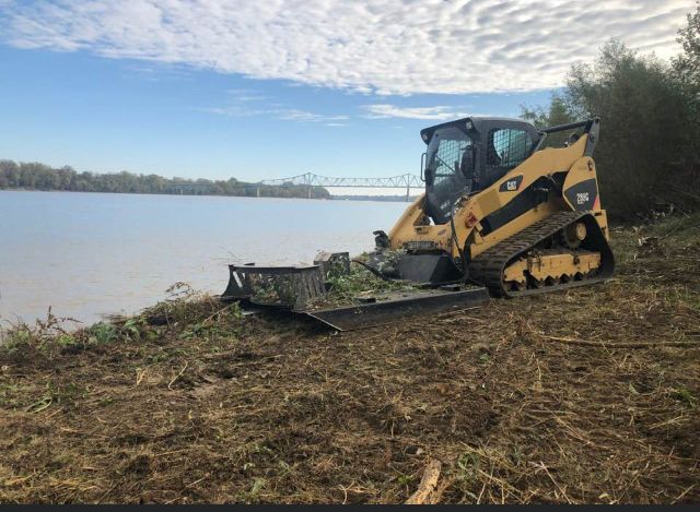 A yellow Caterpillar skid steer with a forestry cutter attachment clearing brush along a riverbank.