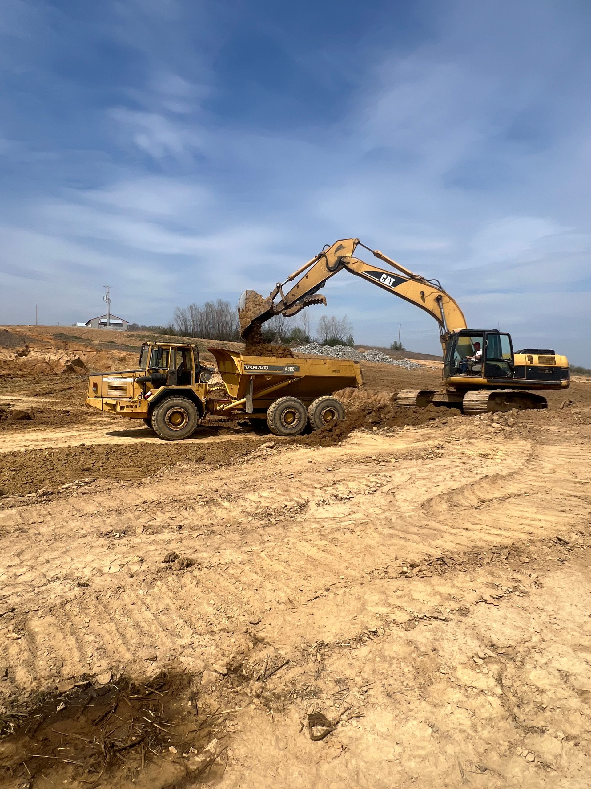 A yellow excavator fills a large, yellow articulated dump truck with dirt at a construction site under a clear blue sky.