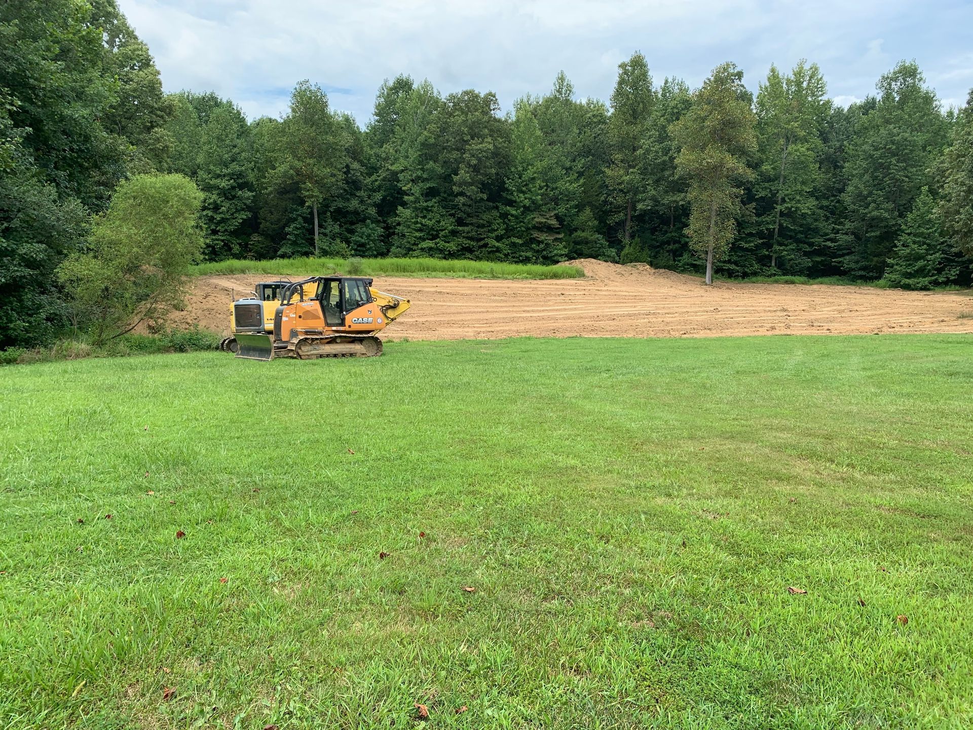 A yellow bulldozer sits on a dirt plot bordering a green field and a line of dense trees under a cloudy sky.