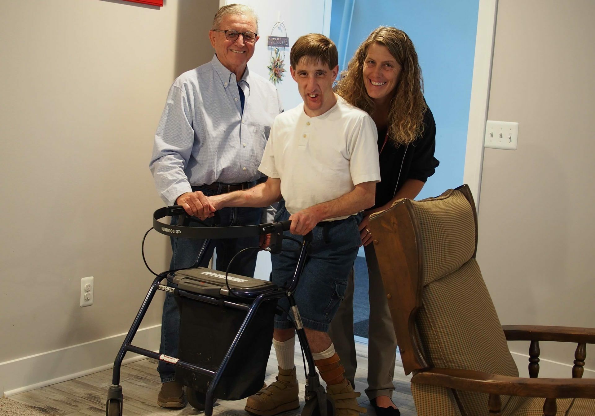 A man with a walker is standing next to two people and a chair.