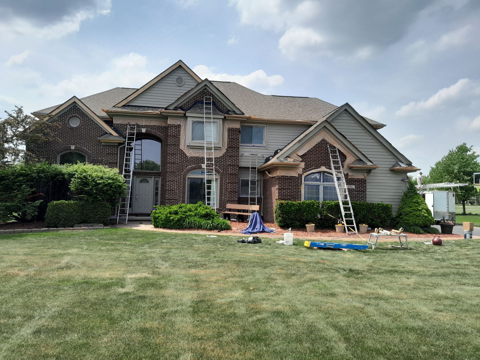 A large brick house is being painted with a ladder in front of it.