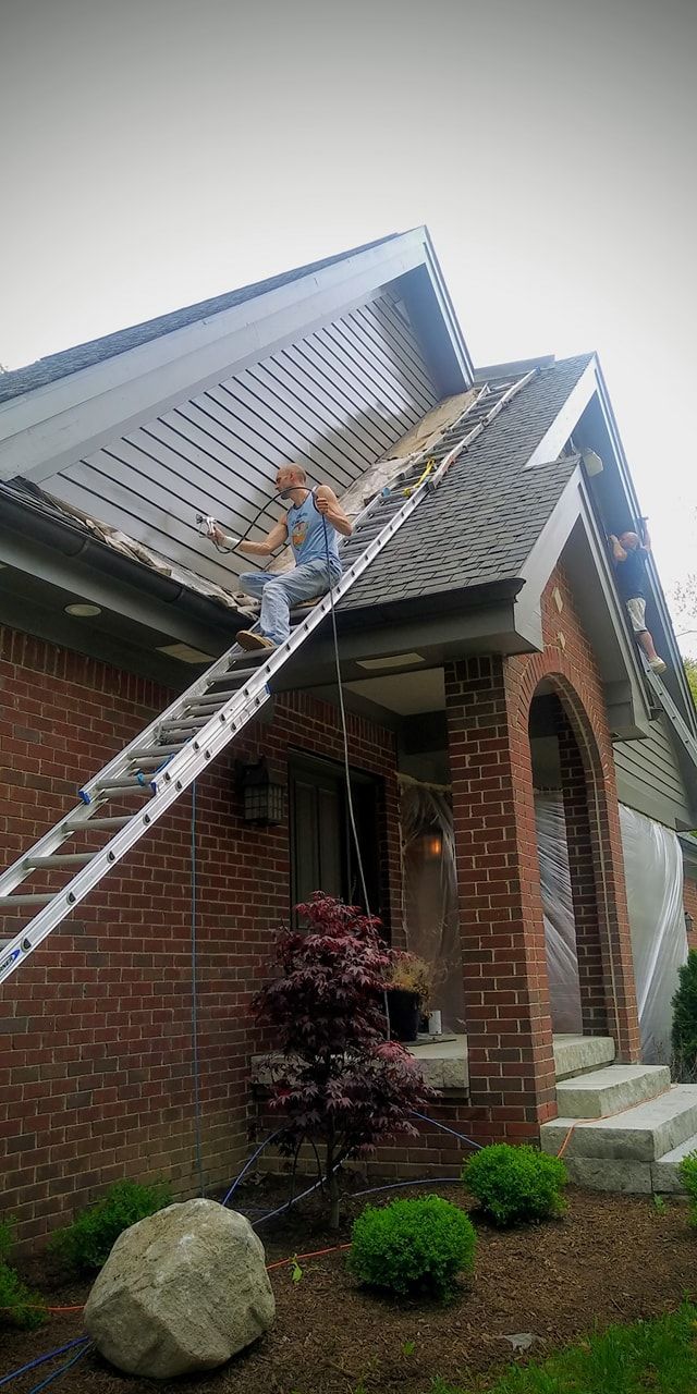 A man is standing on a ladder on the roof of a brick house.
