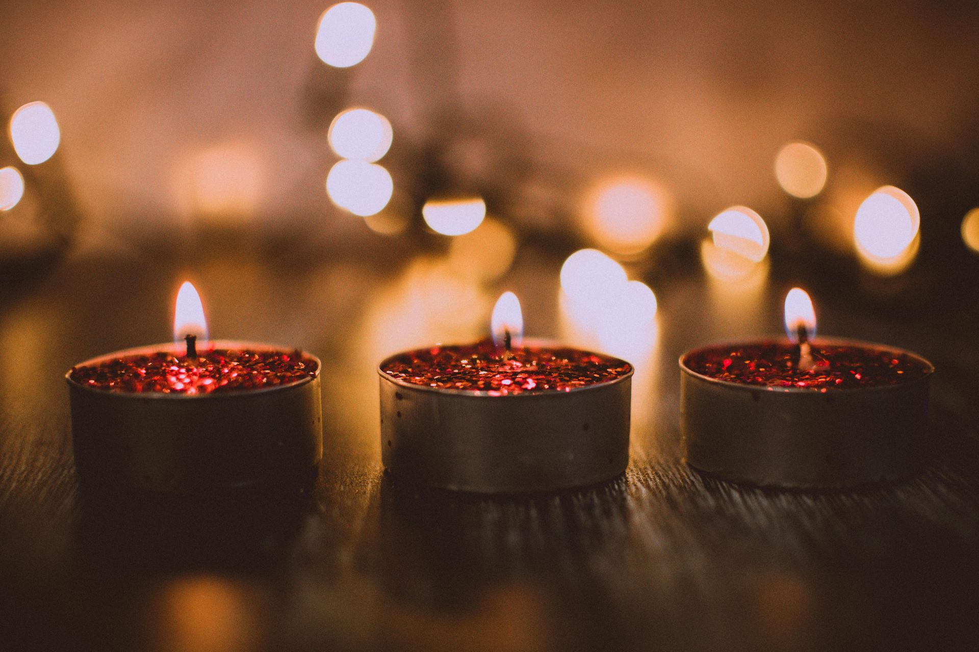 Three lit candles glowing on a dark table with warm, blurred lights in the background