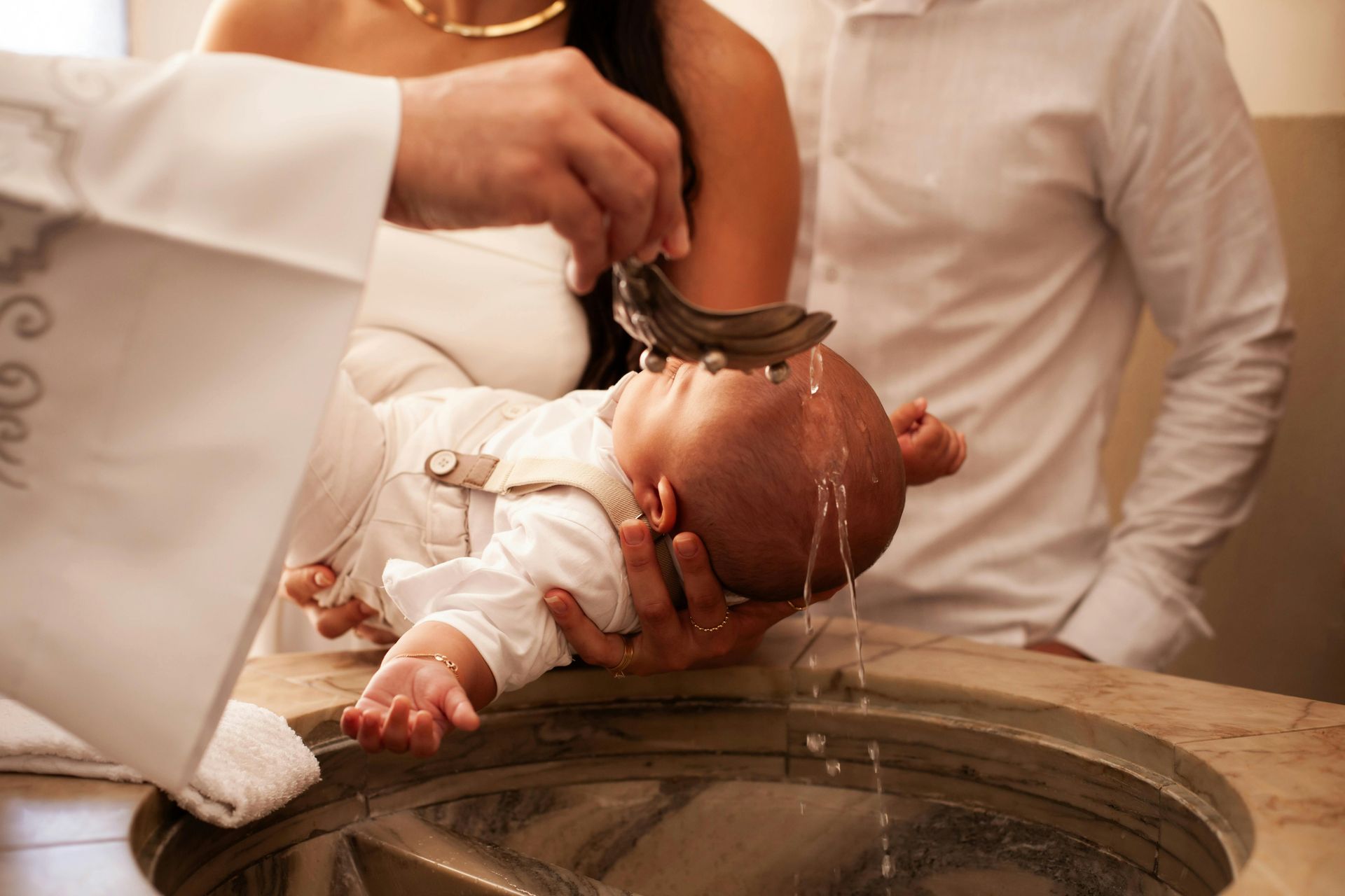 Baby being baptized with water over a stone font, held by an adult in white robes