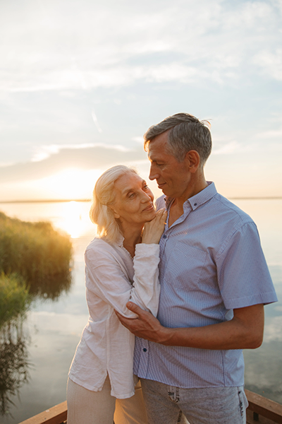 an elderly couple is standing next to each other on a pier near a lake at sunset .