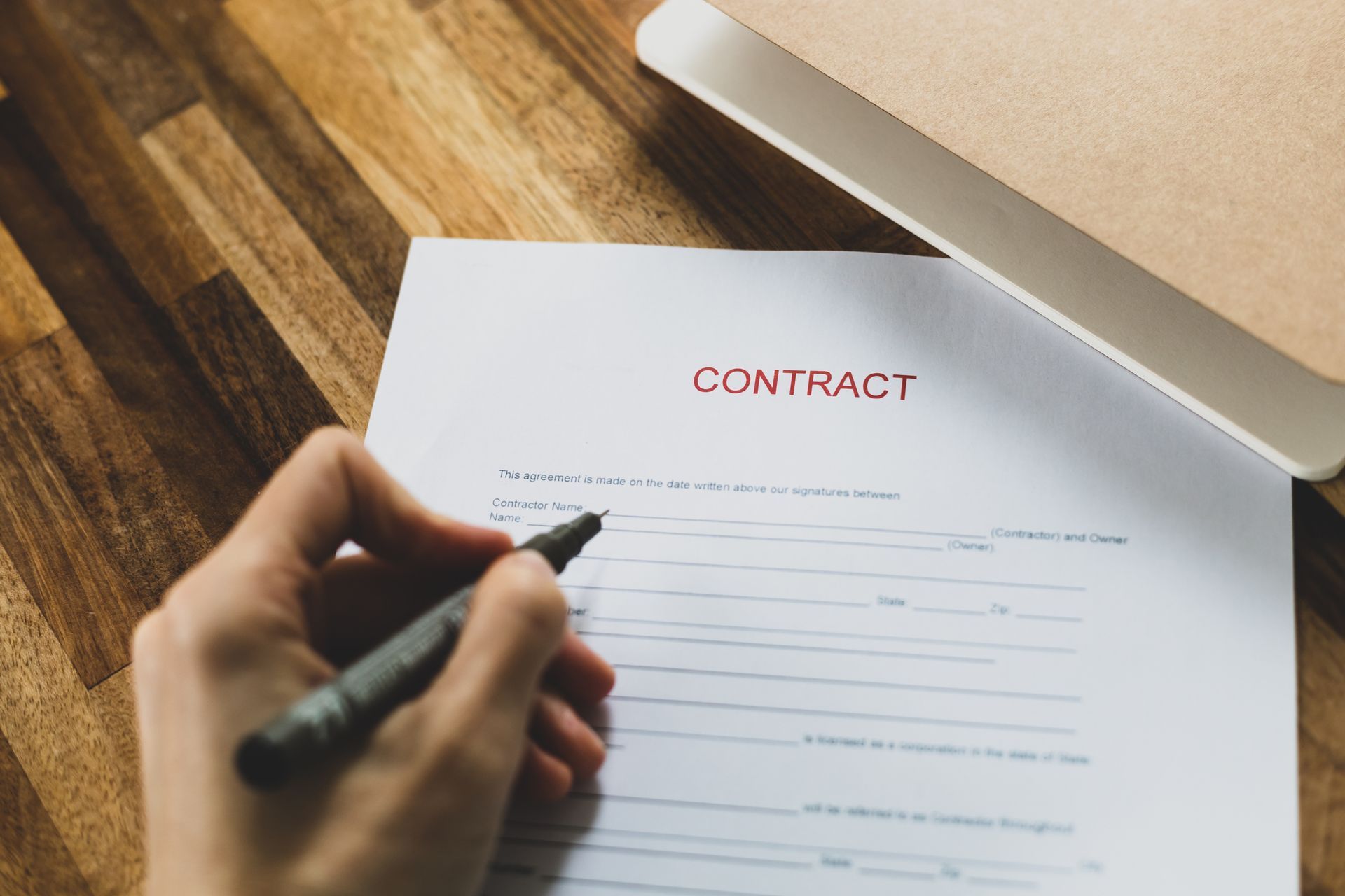 Hand signing a contract with a pen on a wooden table, next to a closed notebook.